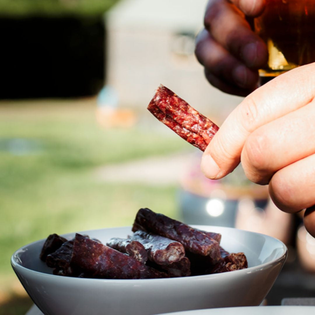 A close-up of a hand holding a piece of drywors against a sunny outdoor backdrop, with a bowl full of these traditional South African dried sausages in the foreground. The setting suggests a relaxed, al fresco dining experience, possibly a barbecue or picnic, with the focus on the rustic, flavourful snack.