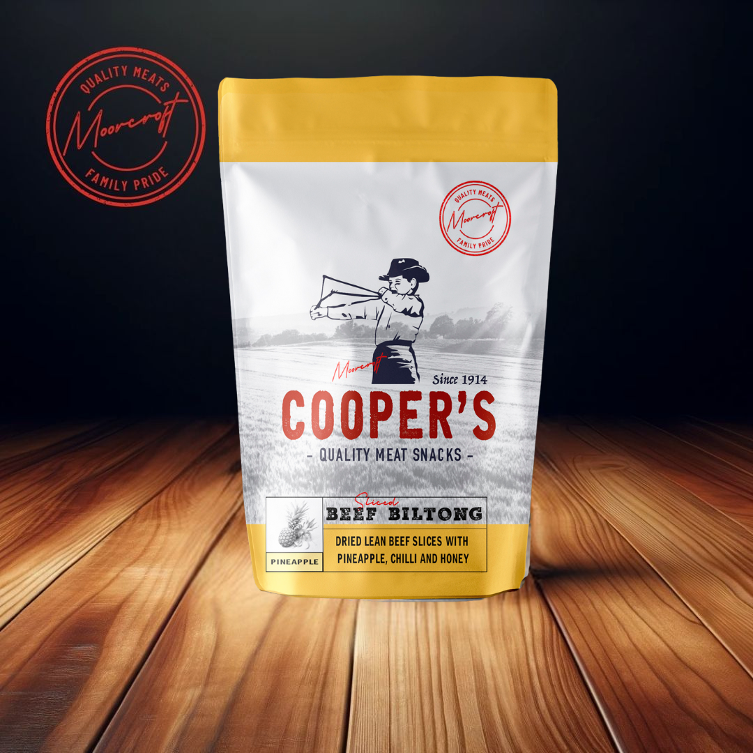 On a polished wooden surface, a pouch of Cooper's Beef Biltong with a distinctive yellow top stands out, offering a unique blend of dried lean beef slices infused with pineapple, chilli, and honey. The packaging proudly displays the Moorcroft Family Pride seal, signifying quality meat snacks with a British heritage since 1914. This gourmet flavour combination suggests a sweet and spicy taste experience