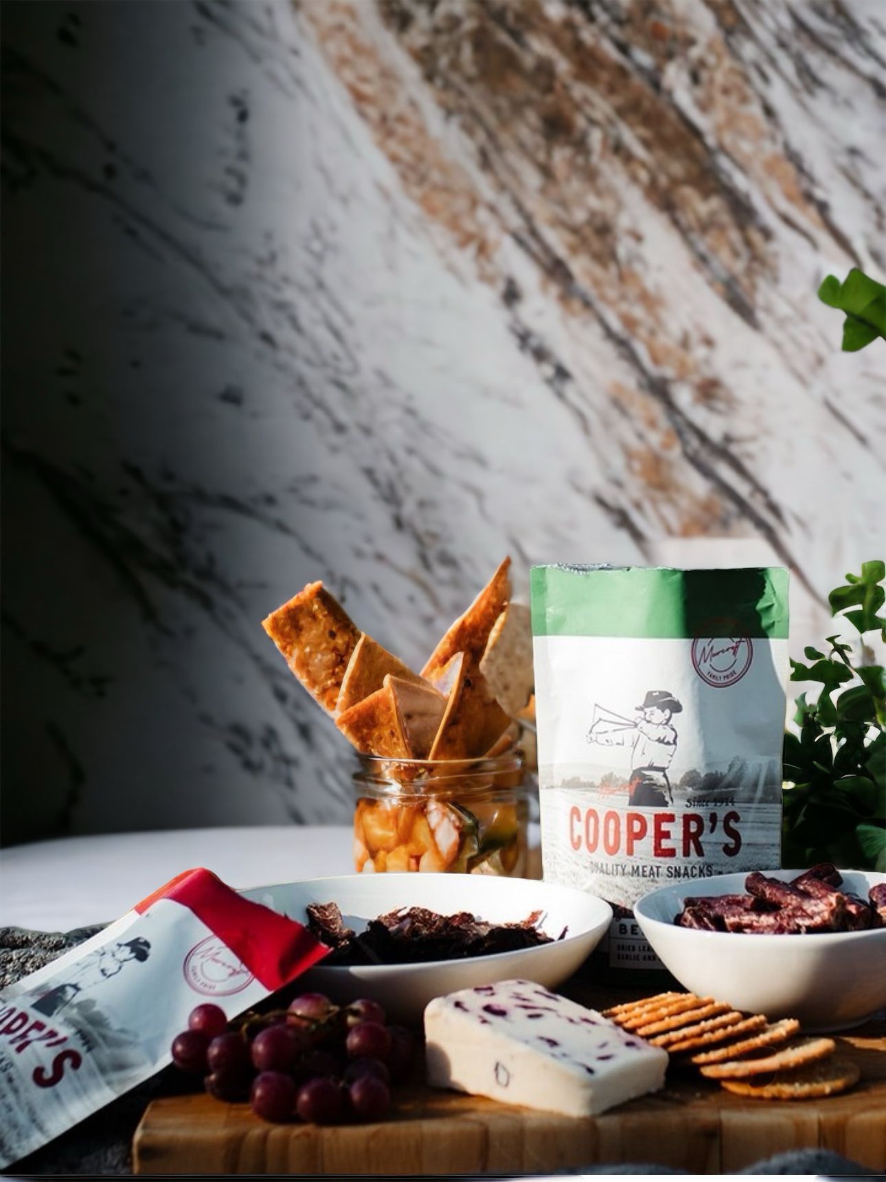 A variety of biltong snacks are prominently displayed, with a bowl of sliced biltong and a packet of Cooper's biltong against a marble backdrop. Accompanying the biltong are a jar of pickled vegetables, fresh grapes, and a selection of crackers and cheese, arranged on a rustic wooden board for an appealing snack presentation.