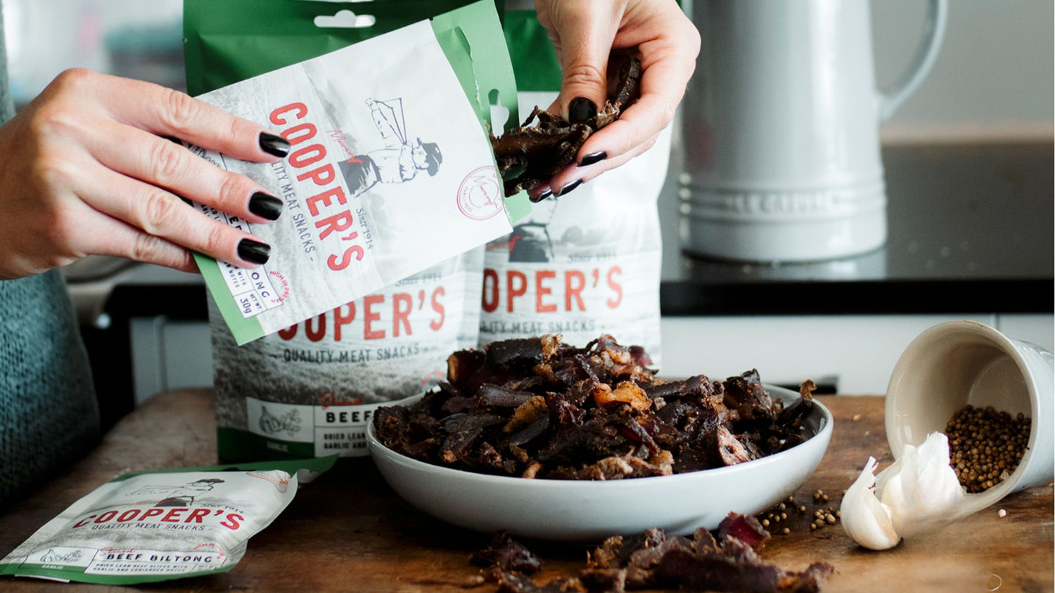 A pair of hands, adorned with dark polished nails, is captured delicately selecting pieces of Cooper's Beef Biltong from a package, adding to a bowlful of the savoury snack on a wooden kitchen worktop. Surrounding the scene are additional packets of Cooper's Biltong, a classic kettle, and culinary essentials such as coriander seeds and garlic, all contributing to a homely British kitchen setting.