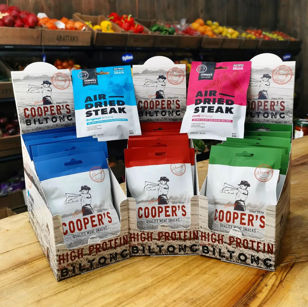 A display of Cooper's High Protein Biltong packets arranged in a wooden stand at a market, with Fenner's Air Dried Steak packets interspersed. The packets are colour-coded—blue for Original and pink for Chilli—prominently placed against a backdrop of fresh produce. The stand invites passersby to try these quality meat snacks, showcasing a variety of flavours.