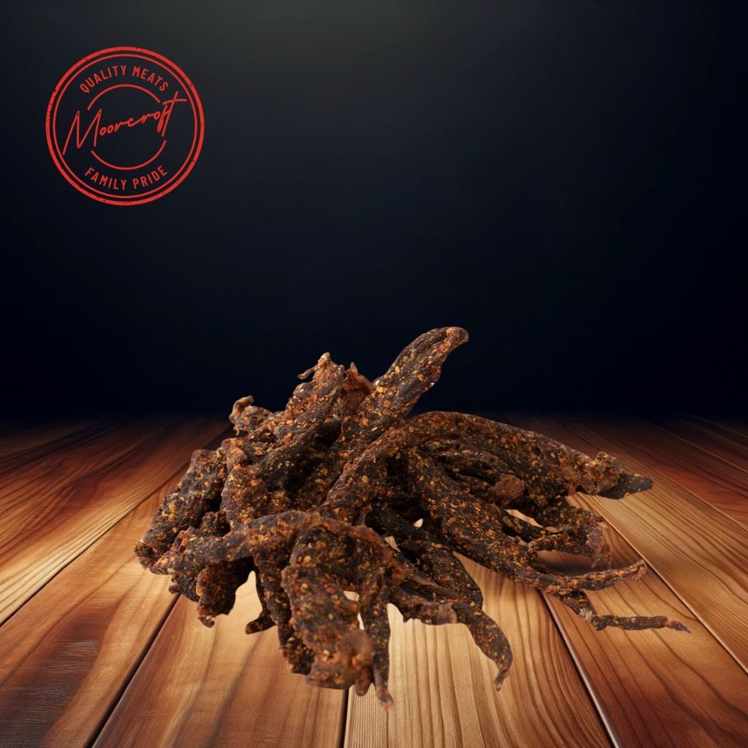 A heap of Cooper’s Chilli Stix biltong scattered across a wooden surface, with a rich, dark background enhancing the textures of the spice-coated meat snacks. The sticks are dark in color, indicating a savory, spicy flavor profile, and the Moorcroft Family Pride seal is visible above, signifying the brand's commitment to quality meat snacks.