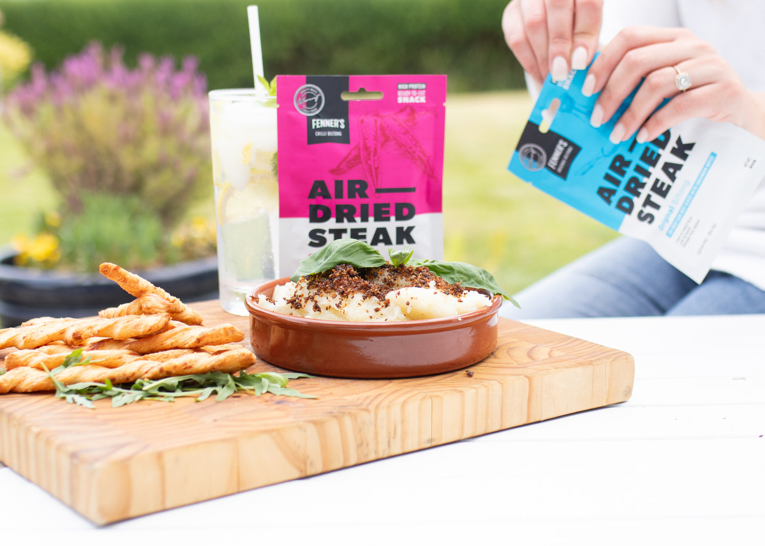 An outdoor snack setup featuring air-dried steak snacks from Fenner's, with a person's hand holding a blue packet on the right and a pink packet standing on the wooden serving board. The board also holds a bowl of mashed potatoes topped with herbs, a stack of twisted breadsticks, and a refreshing drink with a straw, all against a garden background with blooming lavender