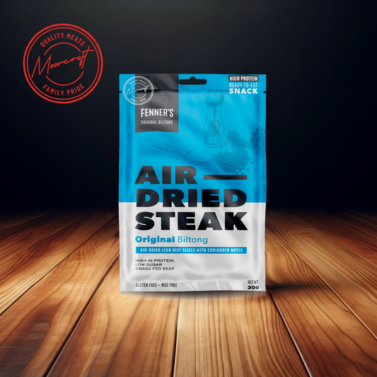 A package of Fenner's Air Dried Steak Original Biltong is prominently displayed on a wooden table, with a cool blue gradient design indicating a high-protein, ready-to-eat snack. The product features air-dried lean beef slices with coriander notes, advertised as high in protein, low in sugar, grass-fed beef, gluten-free, and MSG-free. The net weight is specified as 30g, and the Moorcroft Family Pride emblem assures the quality of the British meat snack