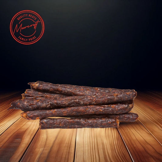 A neat stack of Cooper’s Drywors on a wooden table, the sticks embody the rich tradition of South African cured meat. The original and garlic flavors are implied by their appetizing appearance, and the Moorcroft Family Pride emblem hovers above, suggesting a guarantee of quality. The dark backdrop serves to highlight the artisanal texture and deep color of the Drywors, inviting a taste of these savoury snacks.