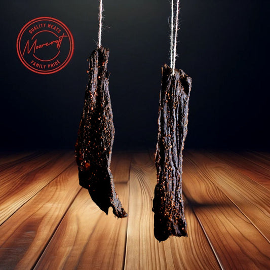 Two thick slabs of Fenner’s Biltong, each weighing 500g, are suspended by strings above a wooden surface, evoking the traditional air-drying process. The richly darkened meat is encrusted with spices, indicative of the slab's robust flavour. In the background, the Moorcroft Family Pride seal affirms the high quality of these British artisanal meat snacks