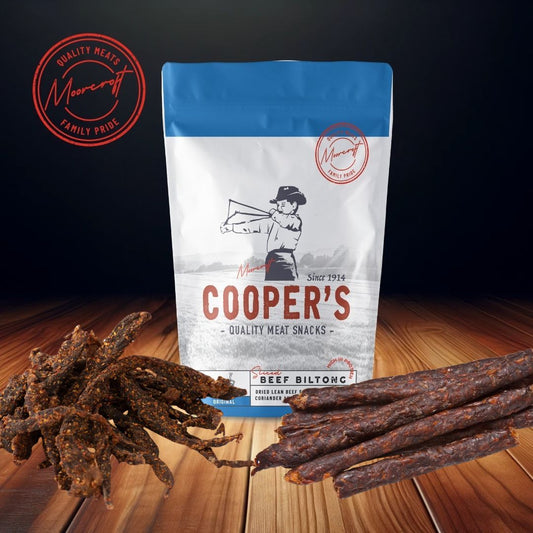 An appetising display of Cooper's biltong, including a packet of the original flavour adorned with the Moorcroft Family Pride seal, alongside a generous portion of chilli stix and drywors laid out on a wooden surface. The packaging's blue and white colouring contrasts with the rich, spiced hues of the meat snacks, all set against a dark background that draws focus to the savoury selection.
