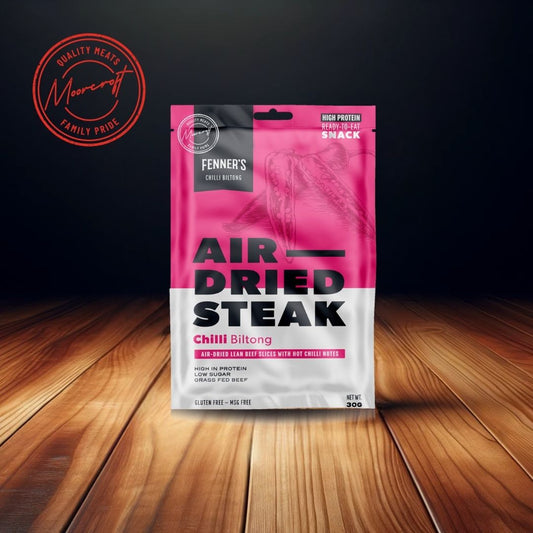 A packet of Fenner's Air Dried Steak Chilli Biltong rests on a wooden surface against a dark background. The packaging is vibrant with a gradient of pink to white, highlighting the product as a high-protein, ready-to-eat snack. It promises air-dried lean beef slices with hot chilli notes, grass-fed beef quality, and is marked as gluten-free and MSG-free, weighing 30g