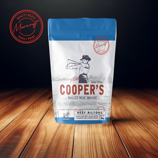 A stand-up pouch of Cooper's Original Beef Biltong is centrally positioned on a wooden table, with a rich, dark background that accentuates the product's classic branding. The package features a distinguished blue top and bears the Moorcroft Family Pride seal, hinting at a longstanding British heritage since 1914. The biltong is described as dried lean beef slices with coriander and pepper notes, offered in a 30g portion.