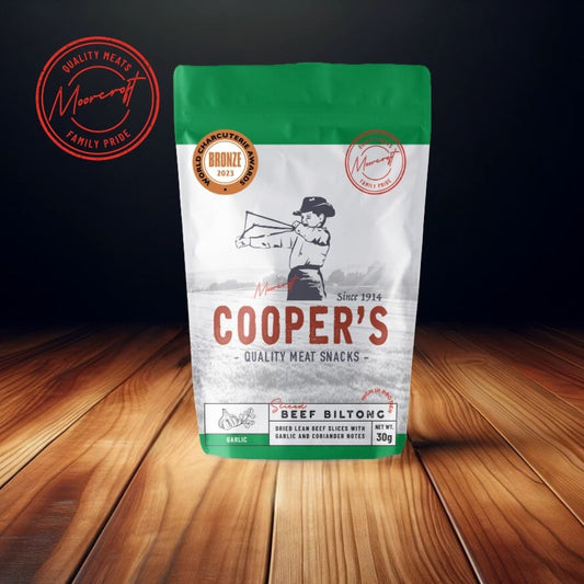 On a polished wooden surface, a packet of Cooper's Garlic Beef Biltong stands out with its green and white colour scheme and 'Bronze 2023' charcuterie award emblem. The product, boasting the Moorcroft Family Pride seal, reflects a longstanding tradition since 1914. The dark backdrop accentuates the biltong's esteemed quality in this British culinary presentation.