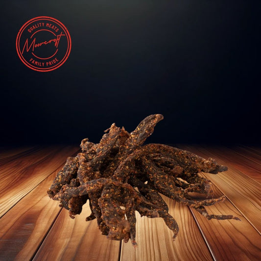 A heap of Cooper’s Chilli Stix biltong scattered across a wooden surface, with a rich, dark background enhancing the textures of the spice-coated meat snacks. The sticks are dark in color, indicating a savory, spicy flavor profile, and the Moorcroft Family Pride seal is visible above, signifying the brand's commitment to quality meat snacks.